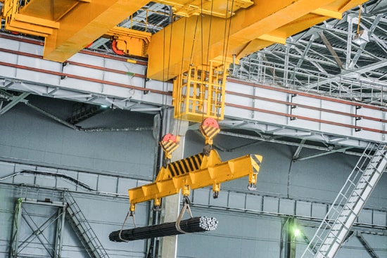 A large double girder crane carries a load of steel rods hung from straps