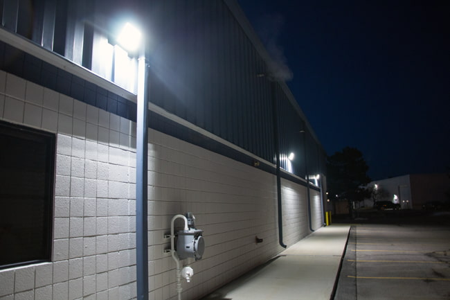 An array of LED wall packs mounted on an exterior wall of a commercial building illuminate an adjacent walkway with parking spaces.