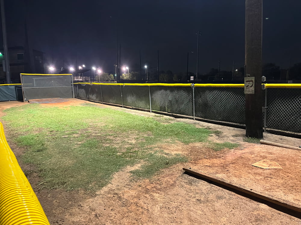The area around an outdoor batting cage and pitching cage with bright illumination from LED flood and sports lights on poles.