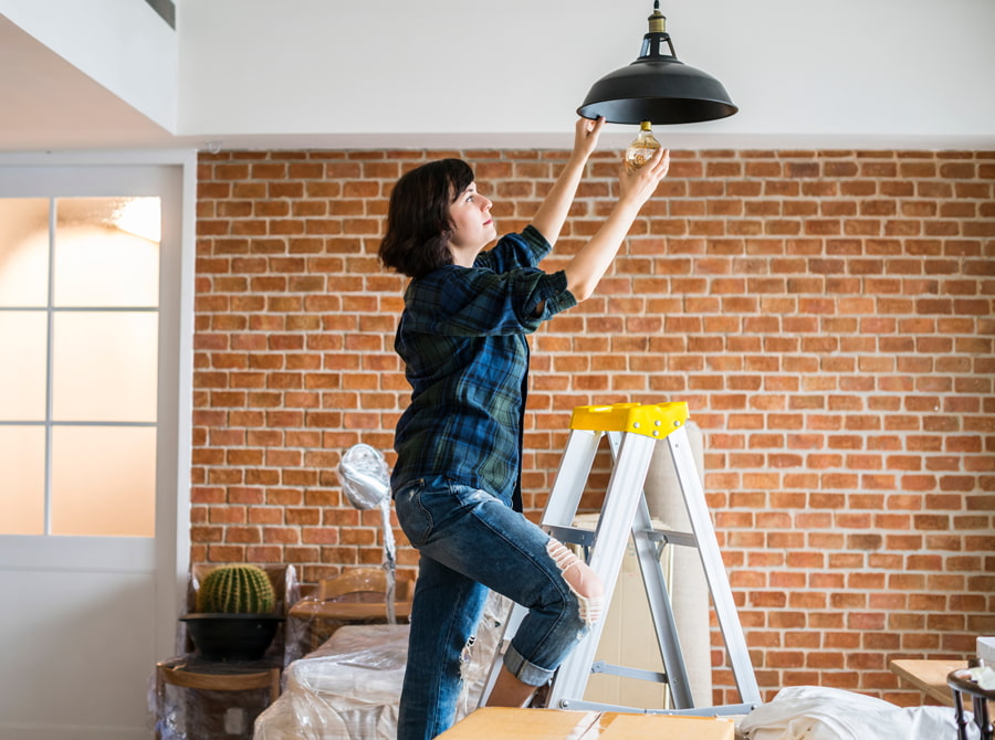 Woman in a plaid shirt and ripped jeans installing a light bulb in a hanging lamp shade