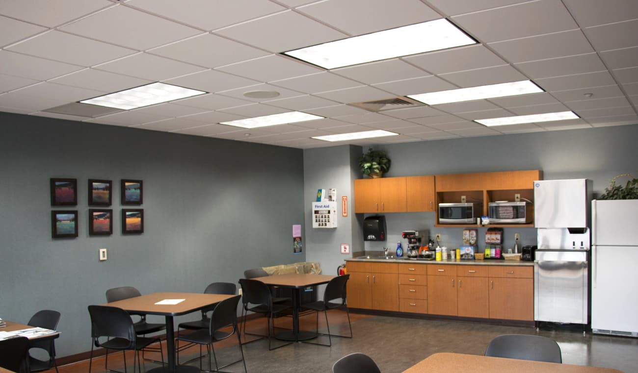 A corporate break room complete with a kitchen is illuminated using LED troffer indoor lighting fixtures
