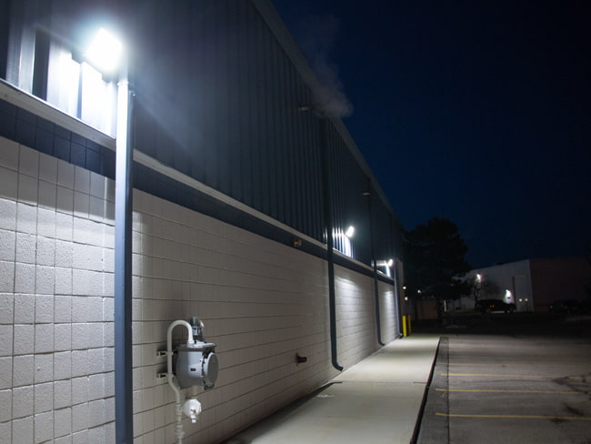 LED Wall packs mounted high above provide exterior building lighting for a commercial facility. They illuminate the sidewalk as well as the parking lot below.