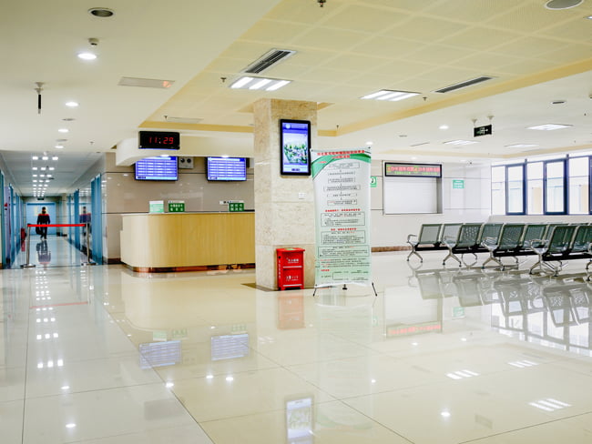 A hospital waiting room with multiple rows of chairs facing the main check in desk and a hallway to the left. There are several round and square shaped hospital lighting fixtures on the ceiling that are illuminating the area.