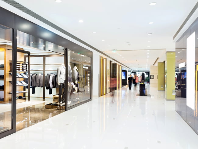 The main corridor of an indoor shopping mall is shown with recessed LED lighting fixtures illuminating retail shops and walking areas