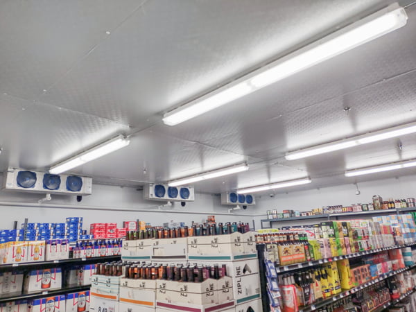A walk in cooler is shown with multiple rows of vapor tight fixtures mounted from the ceiling, illuminating a large beer selection below