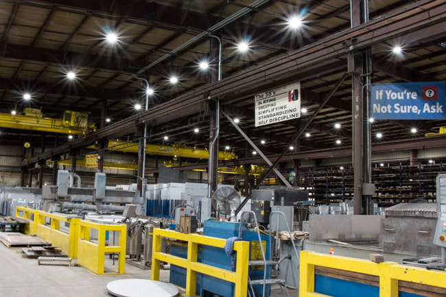 A large metal manufacturing facility is shown with multiple LED high bay lights suspended above providing intense illumination to the shop floor and heavy equipment below