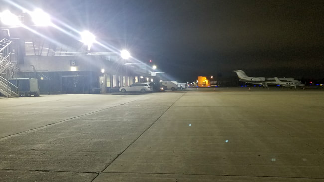 High powered LED flood lights mounted on the sides of aircraft maintenance facilities next to the airports apron area. These lights are clearly illuminating the area for ground crews.