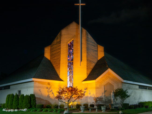 Flood lights illuminate a crucifix on a church with stained glass windows. Wall pack lighting is clearly shown to either side illuminating landscaping.